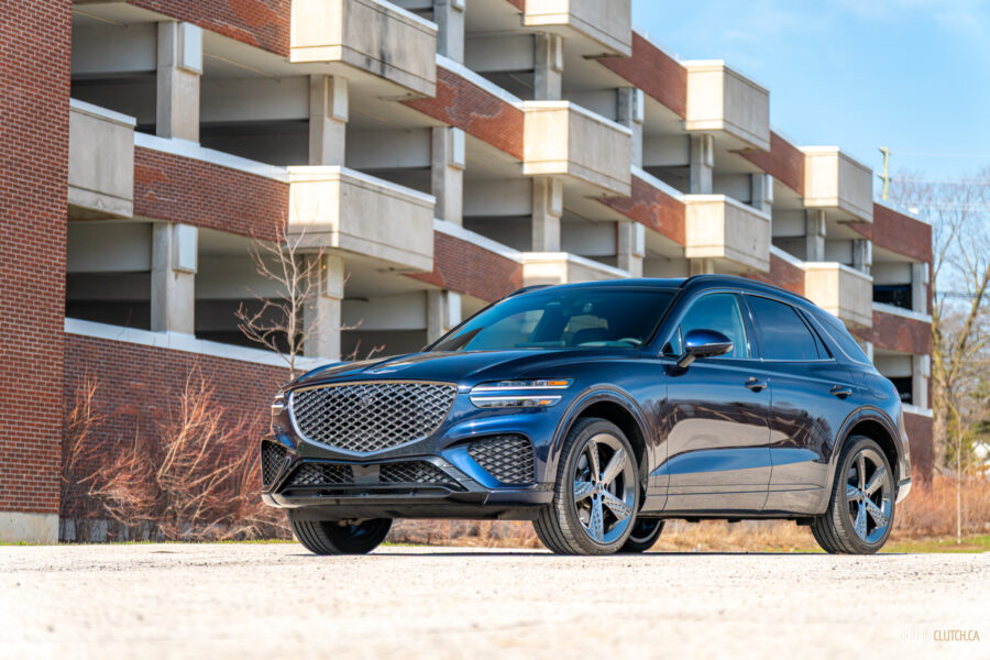 Front quarter view of a dark blue 2025 Genesis GV70 3.5T Sport Plus on a sunny day, with a building in the background