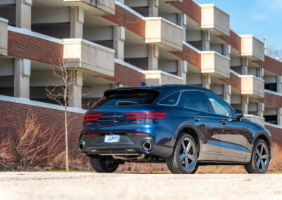 Rear quarter view of a dark blue 2025 Genesis GV70 3.5T Sport Plus on a sunny day, with a building in the background