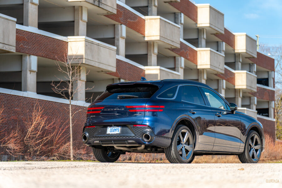 Rear quarter view of a dark blue 2025 Genesis GV70 3.5T Sport Plus on a sunny day, with a building in the background