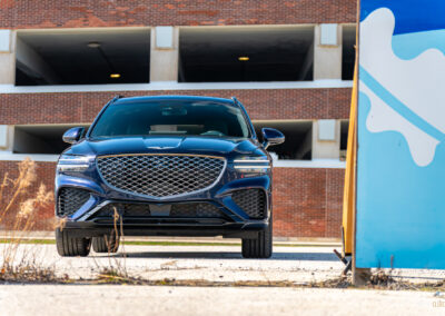Front view of a dark blue 2025 Genesis GV70 3.5T Sport Plus on a sunny day, with a building in the background
