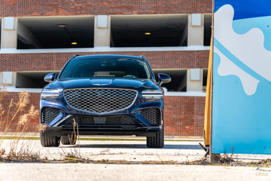 Front view of a dark blue 2025 Genesis GV70 3.5T Sport Plus on a sunny day, with a building in the background