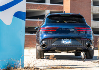 Rear view of a dark blue 2025 Genesis GV70 3.5T Sport Plus on a sunny day, with a building in the background