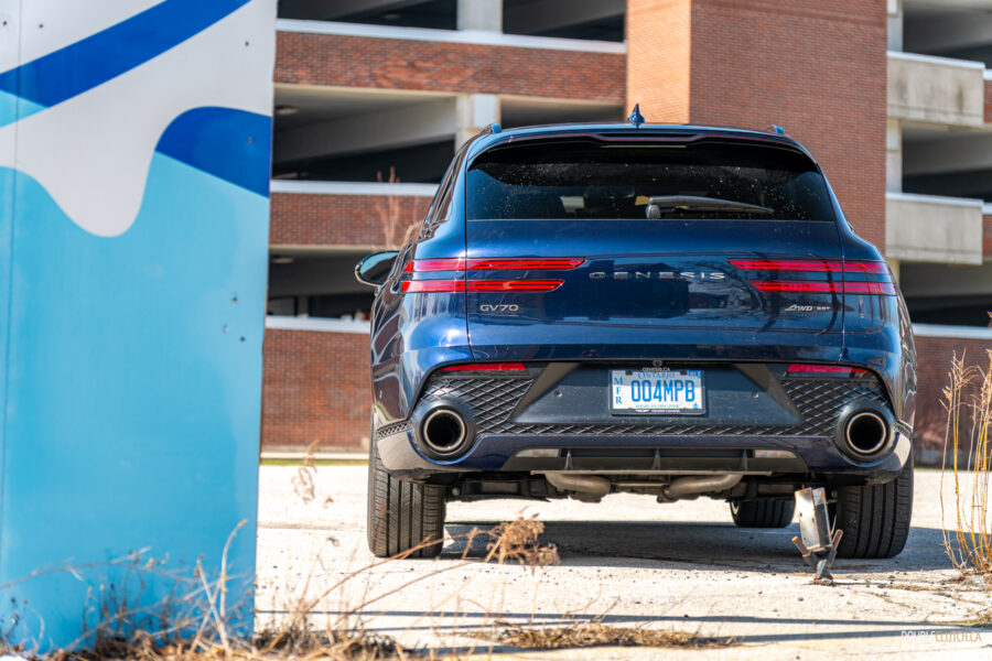 Rear view of a dark blue 2025 Genesis GV70 3.5T Sport Plus on a sunny day, with a building in the background