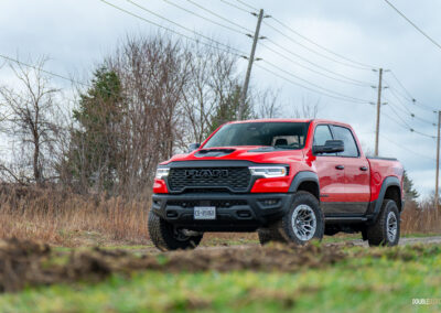 Front quarter view of a red 2025 Ram 1500 RHO on a cloudy day parked on a dirt road, with trees and powerlines in the background