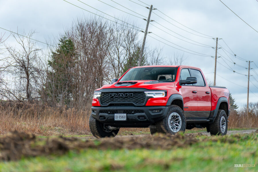 Front quarter view of a red 2025 Ram 1500 RHO on a cloudy day parked on a dirt road, with trees and powerlines in the background