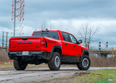 Rear quarter view of a red 2025 Ram 1500 RHO on a cloudy day parked on a dirt road, with trees and powerlines in the background
