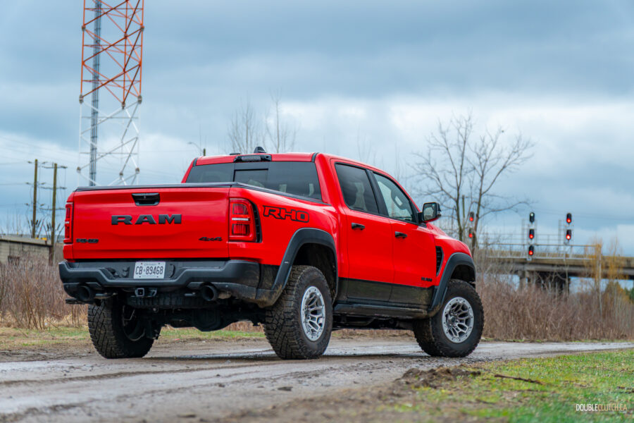Rear quarter view of a red 2025 Ram 1500 RHO on a cloudy day parked on a dirt road, with trees and powerlines in the background