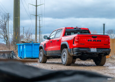 Rear quarter view of a red 2025 Ram 1500 RHO parked in a muddy construction site, with trees and powerlines in the background