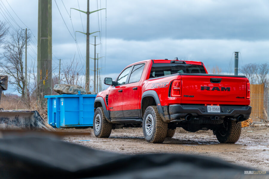 Rear quarter view of a red 2025 Ram 1500 RHO parked in a muddy construction site, with trees and powerlines in the background