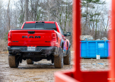 Rear quarter view of a red 2025 Ram 1500 RHO parked in a muddy construction site, with trees and powerlines in the background
