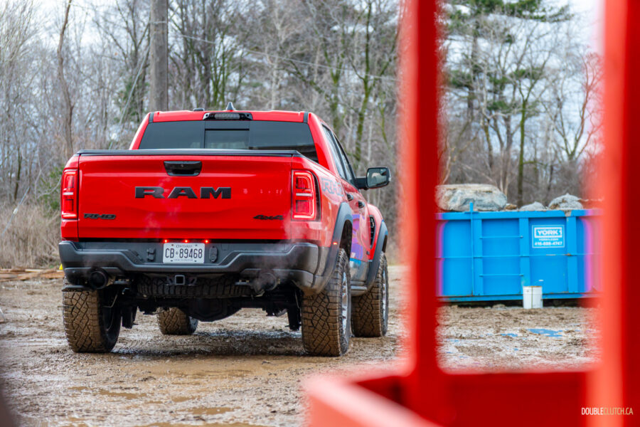 Rear quarter view of a red 2025 Ram 1500 RHO parked in a muddy construction site, with trees and powerlines in the background