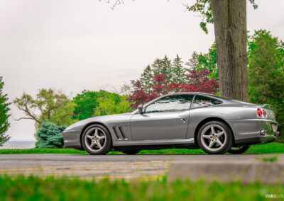 A grey 1996 Ferrari 550 Maranello on a cloudy day, parked on an empty road, with trees, flowers, and shrubs in the background