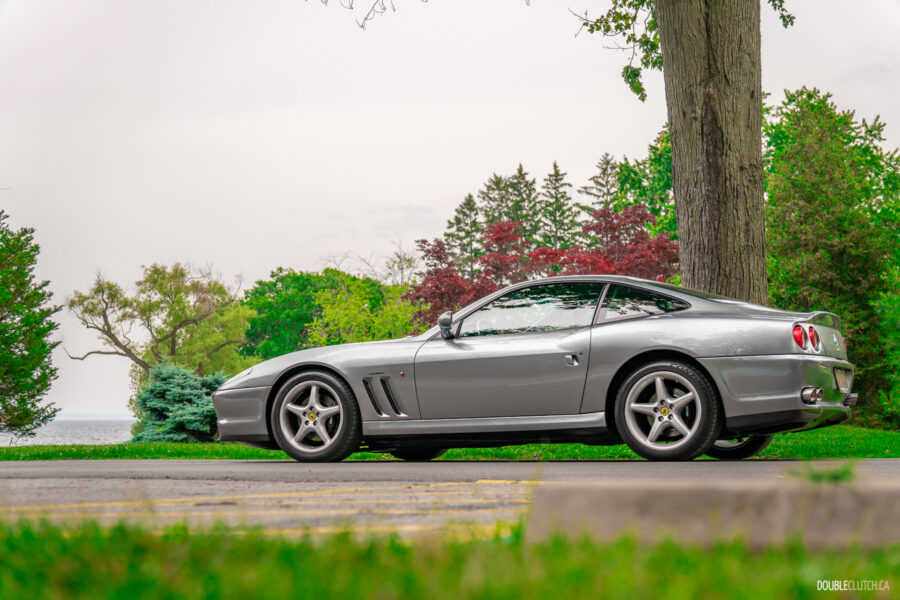 A grey 1996 Ferrari 550 Maranello on a cloudy day, parked on an empty road, with trees, flowers, and shrubs in the background