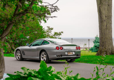 A grey 1996 Ferrari 550 Maranello on a cloudy day, parked on an empty road, with trees and the lake in the background