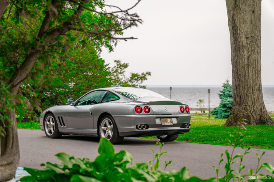 A grey 1996 Ferrari 550 Maranello on a cloudy day, parked on an empty road, with trees and the lake in the background