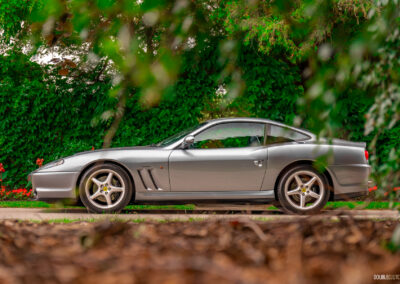 A grey 1996 Ferrari 550 Maranello parked on an empty road, with trees and hedges in the background