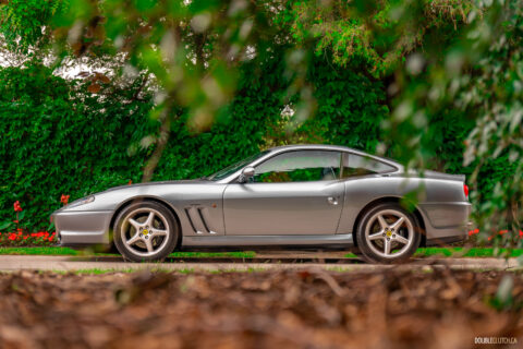 A grey 1996 Ferrari 550 Maranello parked on an empty road, with trees and hedges in the background