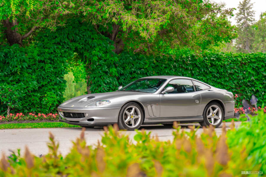 A grey 1996 Ferrari 550 Maranello parked on an empty road, with trees and hedges in the background