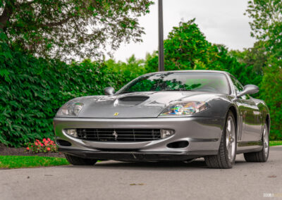 A grey 1996 Ferrari 550 Maranello parked on an empty road, with trees and a lamp post in the background