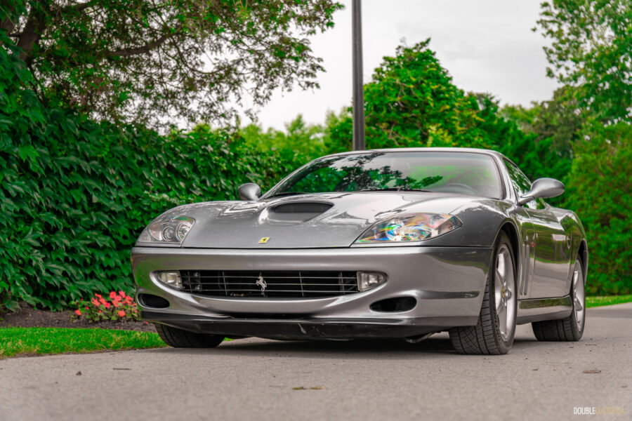 A grey 1996 Ferrari 550 Maranello parked on an empty road, with trees and a lamp post in the background
