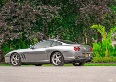 A grey 1996 Ferrari 550 Maranello parked on an empty road, with trees and shrubs in the background