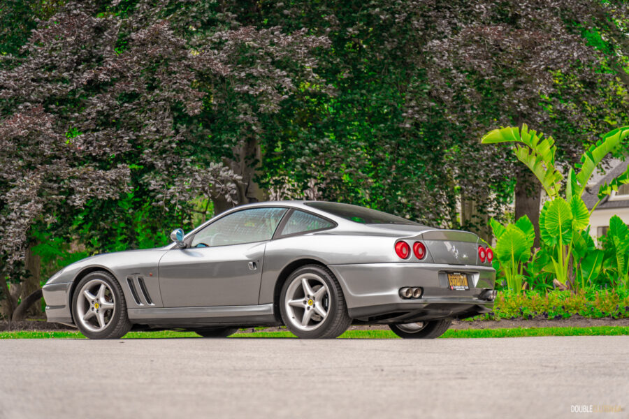 A grey 1996 Ferrari 550 Maranello parked on an empty road, with trees and shrubs in the background