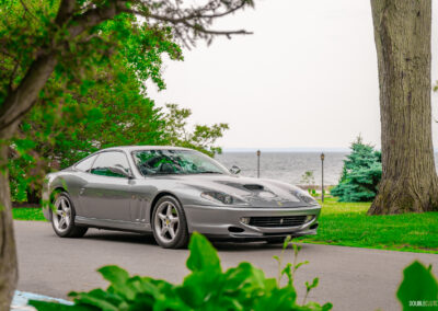 A grey 1996 Ferrari 550 Maranello on a cloudy day, parked on an empty road, with trees and the lake in the background