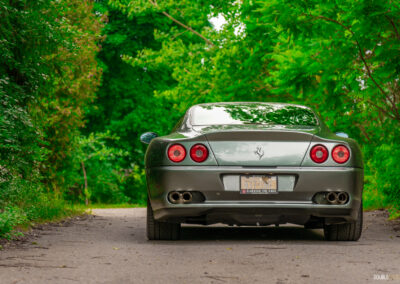 A grey 1996 Ferrari 550 Maranello on a cloudy day, parked on an empty tree-lined road
