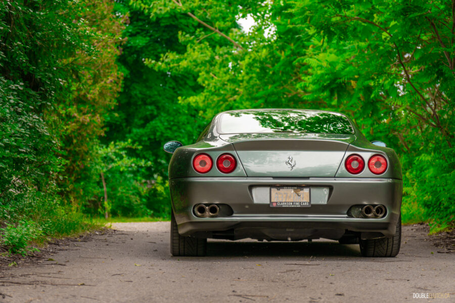 A grey 1996 Ferrari 550 Maranello on a cloudy day, parked on an empty tree-lined road
