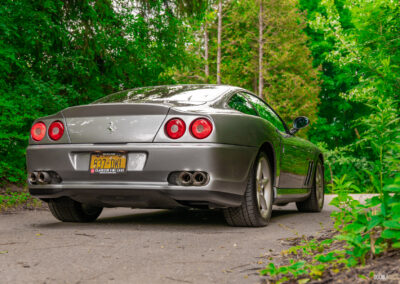 A grey 1996 Ferrari 550 Maranello on a cloudy day, parked on an empty tree-lined road