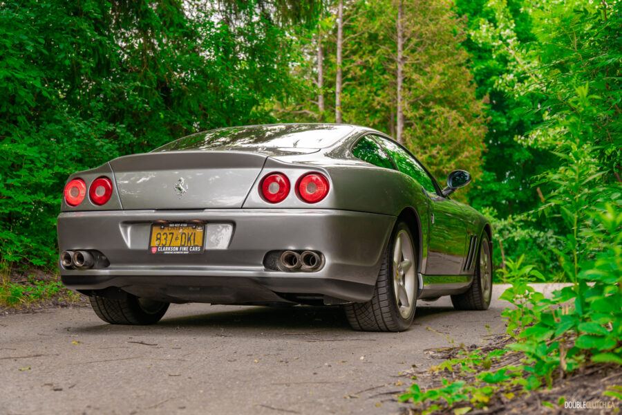 A grey 1996 Ferrari 550 Maranello on a cloudy day, parked on an empty tree-lined road
