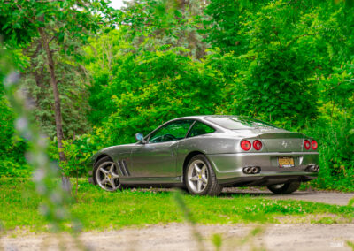 A grey 1996 Ferrari 550 Maranello on a cloudy day, parked on an empty tree-lined road