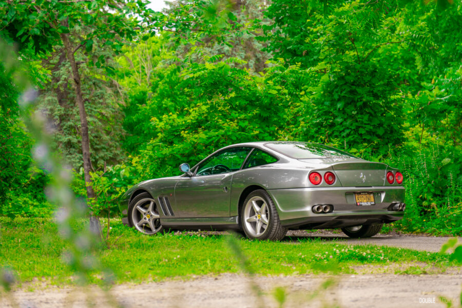 A grey 1996 Ferrari 550 Maranello on a cloudy day, parked on an empty tree-lined road