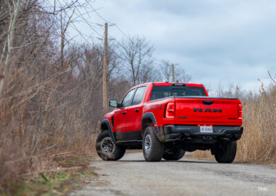 Rear quarter view of a red 2025 Ram 1500 RHO on a cloudy day, with trees and powerlines in the background
