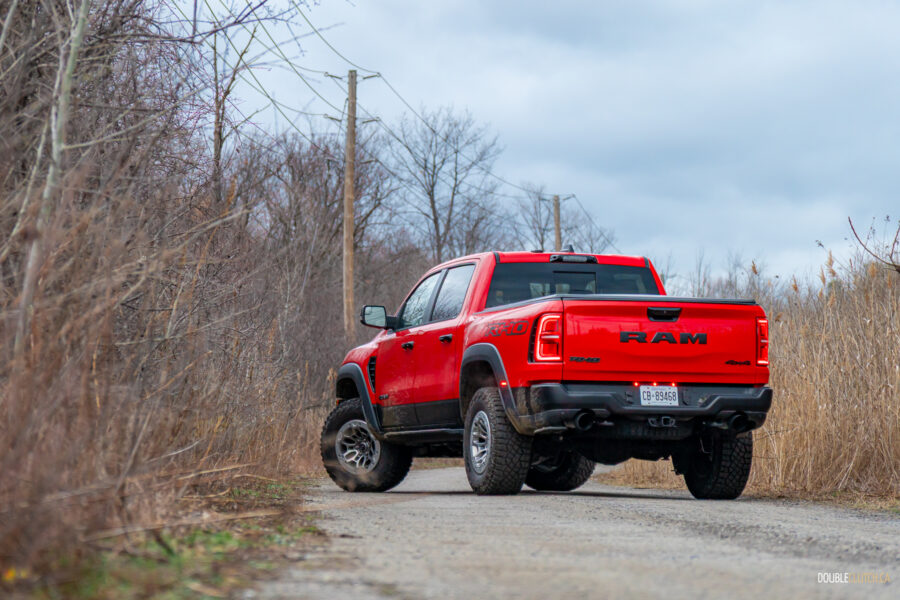 Rear quarter view of a red 2025 Ram 1500 RHO on a cloudy day, with trees and powerlines in the background