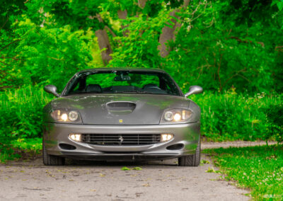 A grey 1996 Ferrari 550 Maranello on a cloudy day, parked on an empty tree-lined road