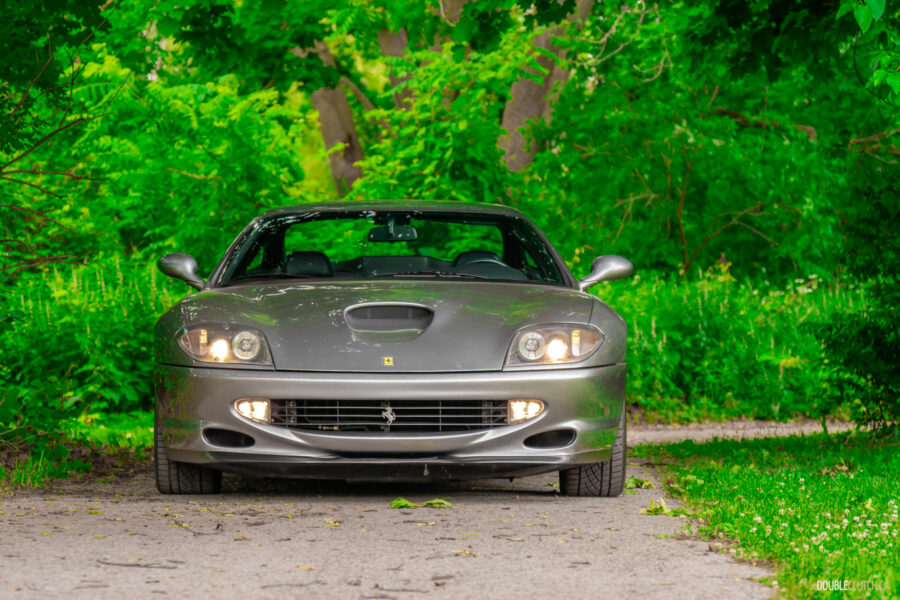 A grey 1996 Ferrari 550 Maranello on a cloudy day, parked on an empty tree-lined road