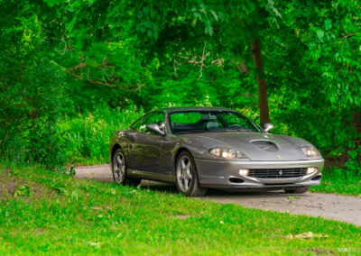A grey 1996 Ferrari 550 Maranello on a cloudy day, parked on an empty tree-lined road