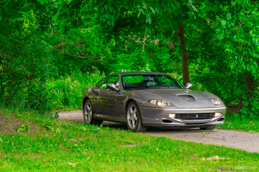 A grey 1996 Ferrari 550 Maranello on a cloudy day, parked on an empty tree-lined road