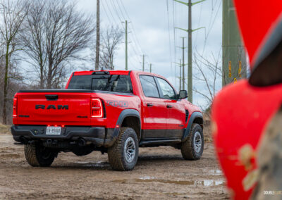 Rear quarter view of a red 2025 Ram 1500 RHO parked in a muddy construction site, with trees and powerlines in the background