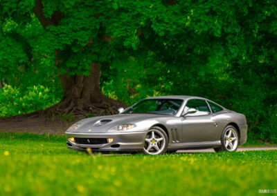 A grey 1996 Ferrari 550 Maranello on a cloudy day, parked on an empty tree-lined road