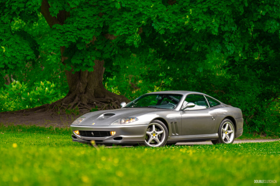 A grey 1996 Ferrari 550 Maranello on a cloudy day, parked on an empty tree-lined road