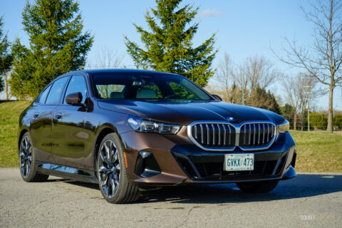 Front quarter view of a brown 2025 BMW 550e parked on an empty road on a sunny day, with trees in the background